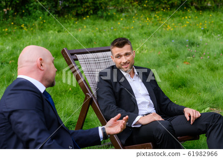 Businessmen are sitting in chairs and talking on grass outdoors 61982966