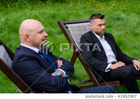 Businessmen are sitting in chairs on grass outdoors Businessmen are sitting in chairs on grass outdoors 61982967