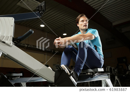 Caucasian male athlete sitting on modern indoor rower and resting after training Caucasian male athlete sitting on modern indoor rower and resting after training 61983887