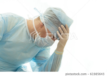 Upset surgeon in sterile gown and surgical mask props his head with hand Upset surgeon in sterile gown and surgical mask props his head with hand 61985643