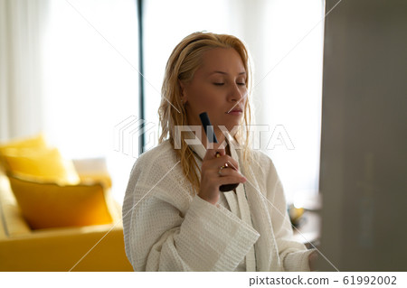 Young beautiful model in a waffle bathrobe gown with closed eyes and a make-up brush in her hands is standing in an apartment with yellow furniture. Young beautiful model in a waffle bathrobe gown with closed eyes and a make-up brush in her hands is standing in an apartment with yellow furniture. 61992002