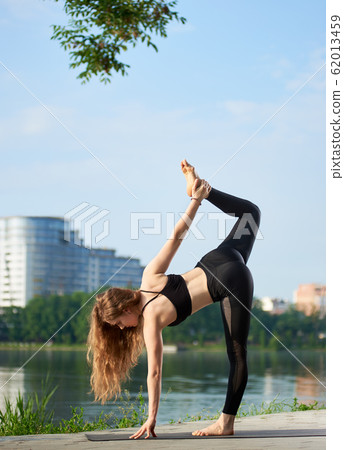 Flexible yoga female with long hair standing in sugarcane pose, ardha chandra chapasana near the river at city park 62013459