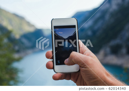 A man takes a picture of mountain landscape on the phone while holding it in one hand A man takes a picture of mountain landscape on the phone while holding it in one hand 62014340