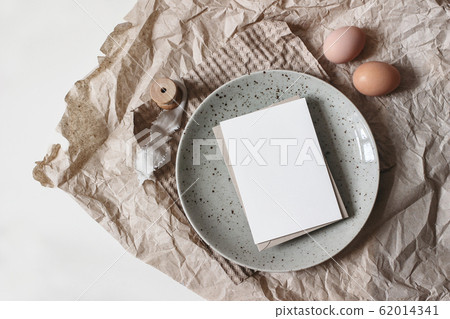 Spring breakfast scene. Lifestyle mockup composition with blank greeting card, ceramic plate and chicken eggs on brown craft paper. White table background. Easter food concept. Flat lay, top view. 62014341