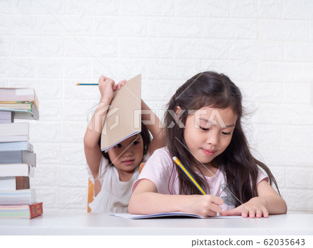 Asian sibling little cute girl sitting and writing at paper on white table with a stack of books. Preschool lovely kid writing homework at home. Learning and education of kid. Asian sibling little cute girl sitting and writing at paper on white table with a stack of books. Preschool lovely kid writing homework at home. Learning and education of kid. 62035643