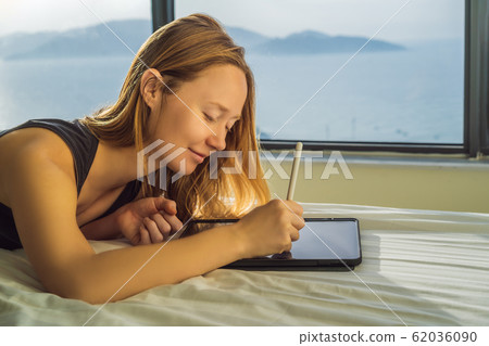 Young woman draws on a tablet against the background of a window with a sea view. Designer Young woman draws on a tablet against the background of a window with a sea view. Designer 62036090