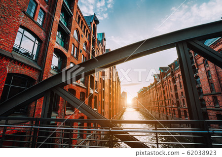 The red brick warehouse - Speicherstadt district in Hamburg Germany, framed by steel bridge arch beams with canal perspective filled by warm sunset light 62038923