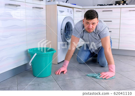 Unhappy tired man in rubber gloves washes the floor in the kitchen and looks at the camera at the end. Unhappy tired man in rubber gloves washes the floor in the kitchen and looks at the camera at the end. 62041110