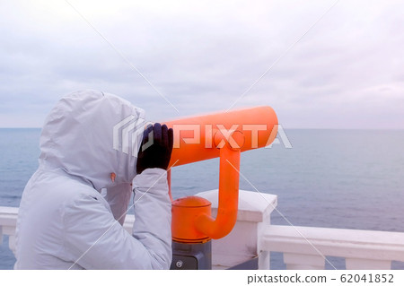 Woman in white jacket and hood looks in binoculars at sea waterfront with sea view. Side view. 62041852