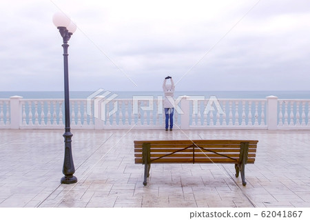 Woman in white jacket and hood on beautiful terrace with sea view on waterfront. Back view. 62041867