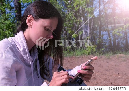Woman is typing message on smartphone sitting on nature, side view. Woman is typing message on smartphone sitting on nature, side view. 62045753