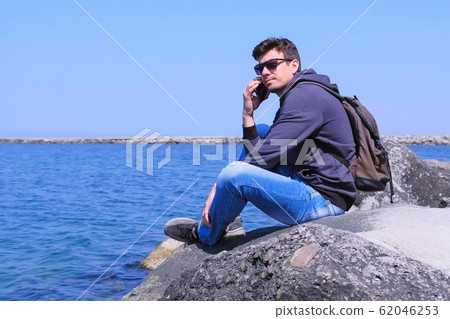 Man traveller calls smartphone sits on stone waits answer on sea background. Man traveller calls smartphone sits on stone waits answer on sea background. 62046253