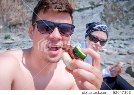 Man blogger is eating shashlik with bread and cucumber on camera with his wife and talking about it his subscribers. 62047308