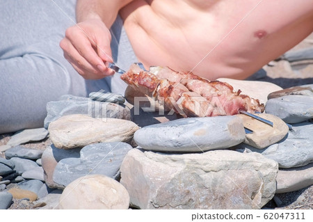 Man is cooking shashlik on campfire rotating skewers lying on the stones, food closeup. 62047311