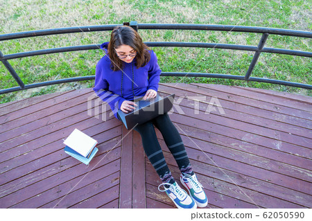 Intellectual young girl sit on the patio and using laptop with books next to her in a park. Intellectual young girl sit on the patio and using laptop with books next to her in a park. 62050590
