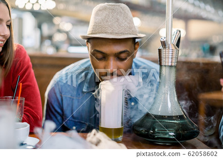 handsome young African man in jeans wear and hat is smoking hookah in a restaurant in daytime, exhaling smoke at beer glass, standing on table 62058602