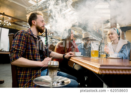 Close-up of Young man in casual wear smoking traditional hookah pipe. man exhaling smoke. Friends in cafe having fun 62058603