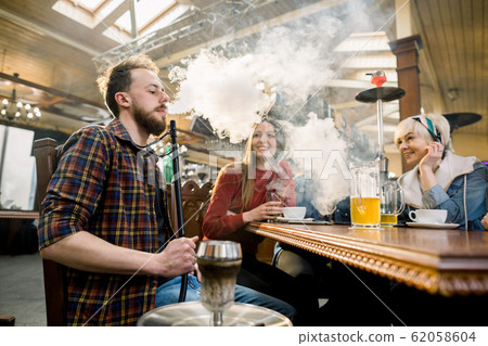 Close-up of Young man in casual wear smoking traditional hookah pipe. man exhaling smoke. Friends in cafe having fun 62058604