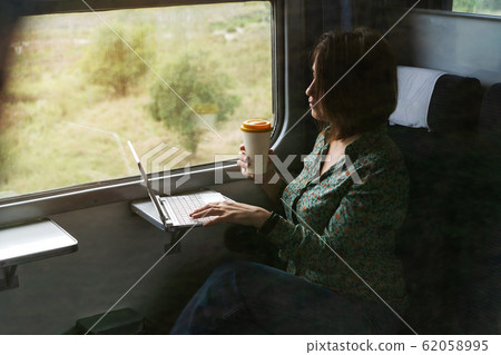 A woman sitting by the window of a commuter train with a laptop and coffee cup 62058995