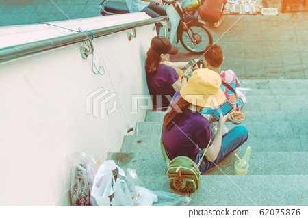 A woman wearing a yellow hat sit to play a smartphone while the other hand hold a cup of meat on the stair in the city 62075876