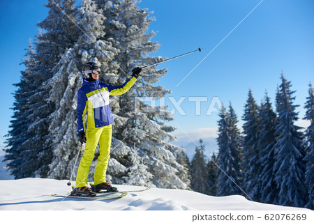 Happy woman posing on skis before skiing. Sunny day at ski resort. Clear blue sky, snow-covered fir trees on background. 62076269