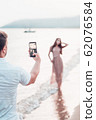 Man taking foto on a the camera on smartphone digital camera of a sporty young beautiful lady on a sea shore background. Outdoor lifestyle picture on a hot sunny summer day. 62076584