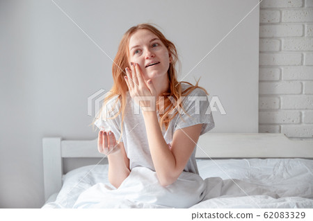 Woman holding bottle of cream with cosmetic products. Blonde woman holding a glass jar with face cream sitting on the bed Woman holding bottle of cream with cosmetic products. Blonde woman holding a glass jar with face cream sitting on the bed 62083329