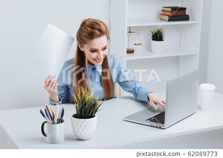 Photo of smiling businesswoman sitting at office and working. Using phone in office. Office worker holding documents. Photo of smiling businesswoman sitting at office and working. Using phone in office. Office worker holding documents. 62089773