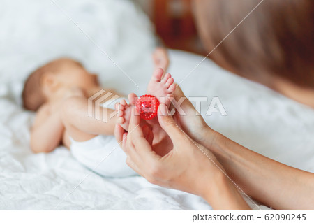 Mother holds newborn baby's feet. Tiny fingers and red massage ball in woman's hand. Cozy morning at home. Mother holds newborn baby's feet. Tiny fingers and red massage ball in woman's hand. Cozy morning at home. 62090245