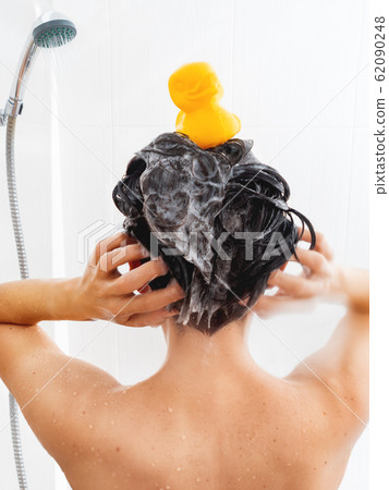 Naked woman with short hair takes a shower. Woman washes her hair with shampoo. Yellow rubber duck on girl's head in white bathroom. Naked woman with short hair takes a shower. Woman washes her hair with shampoo. Yellow rubber duck on girl's head in white bathroom. 62090248