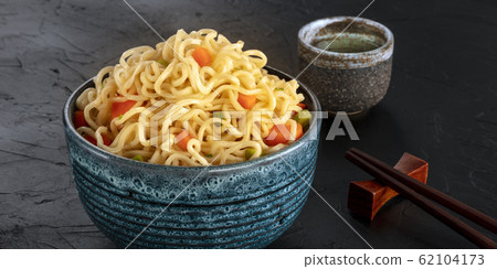 Instant noodles panorama, a bowl of soba with carrot and scallions, with a cup of sake and chopsticks, on a black background Instant noodles panorama, a bowl of soba with carrot and scallions, with a cup of sake and chopsticks, on a black background 62104173