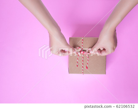 Female hands bow tie a simple craft paper gift box wrapped with colorful rope with as a present for Christmas, new year, valentine day or anniversary on pink background, top view 62106532
