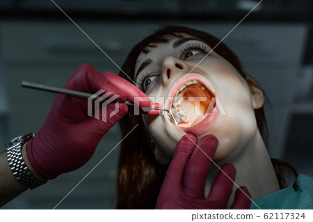 Close up portrait of young female patient with open mouth having professional dental treatment, and hands of doctor wearing red rubber gloves, holding dental mirror 62117324
