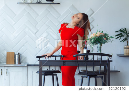 Beautiful girl in a red dress is standing in the kitchen Beautiful girl in a red dress is standing in the kitchen 62120192