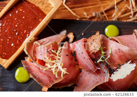 Antipasto on wooden plate close up. Cold smoked meat plate with tomato hot sauce, sliced ham, prosciutto, bacon. Appetizer on wooden tray cut tree sawed imitation. 62126633