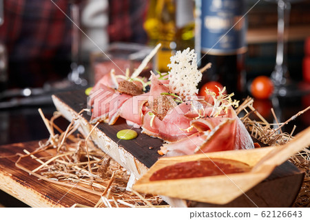 Antipasto on wooden plate close up. Cold smoked meat plate with tomato hot sauce, sliced ham, prosciutto, bacon. Appetizer on wooden tray cut tree sawed imitation. Antipasto on wooden plate close up. Cold smoked meat plate with tomato hot sauce, sliced ham, prosciutto, bacon. Appetizer on wooden tray cut tree sawed imitation. 62126643