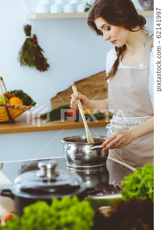 Young brunette woman cooking soup in kitchen. Housewife holding wooden spoon in her hand. Food and health concept Young brunette woman cooking soup in kitchen. Housewife holding wooden spoon in her hand. Food and health concept 62141997