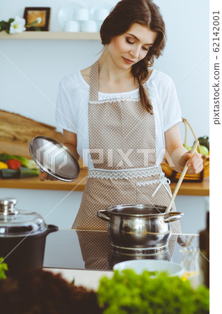 Young brunette woman cooking soup in kitchen. Housewife holding wooden spoon in her hand. Food and health concept 62142001