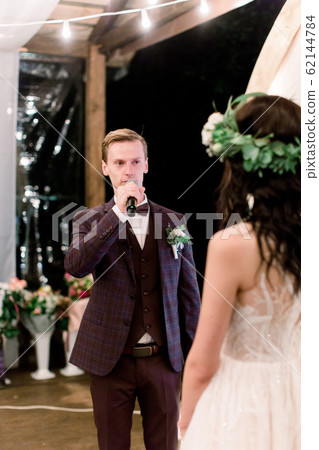 Wedding day. Attractive stylish groom in suit saying an oath, while looking at the bride in elegant rustic dress and wreath on head, during night wedding ceremony. Wooden tent restaurant outdoors 62144784