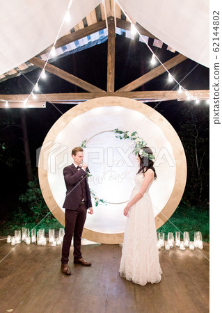 Night wedding ceremony outdoors. Bride and groom swear an oath each other on wedding arch background. Beautiful just married couple. Rustic wooden restaurant with light bulbs 62144802