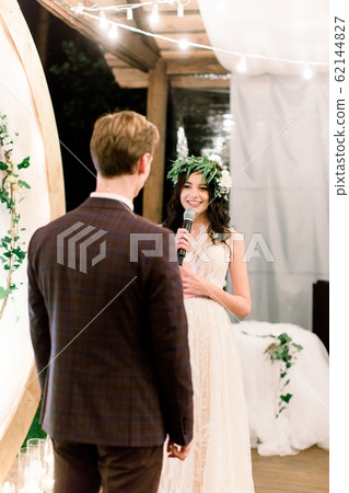 Pretty bride in wedding dress and floral wreath on head is speaking an oath to her groom during night wedding ceremony, standing near the rustic arch. Outdoors. Pretty bride in wedding dress and floral wreath on head is speaking an oath to her groom during night wedding ceremony, standing near the rustic arch. Outdoors. 62144827