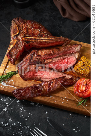 Close up sliced medium rare grilled beef steak with corn and tomato, pepper and rosemary on meat cutting board on dark wooden background Close up sliced medium rare grilled beef steak with corn and tomato, pepper and rosemary on meat cutting board on dark wooden background 62149683