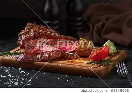 Close up sliced medium rare grilled beef steak with corn and tomato, pepper and rosemary on meat cutting board on dark wooden background 62149688