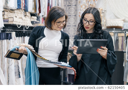 Two women working with interior fabrics digital tablet in showroom for curtains and upholstery fabrics, designer and buyer choosing fabrics in new interior Two women working with interior fabrics digital tablet in showroom for curtains and upholstery fabrics, designer and buyer choosing fabrics in new interior 62150405