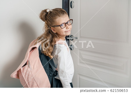 Little pretty girl 6, 7 years old with school backpack. Smiling girl standing near the front door of the house, child goes to school Little pretty girl 6, 7 years old with school backpack. Smiling girl standing near the front door of the house, child goes to school 62150653