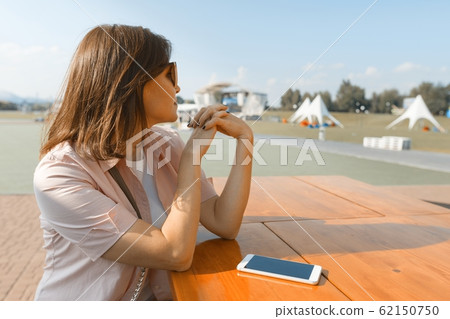Adult middle-aged woman looks away. Background summer street cafe, woman in sunglasses sits at table, copy space Adult middle-aged woman looks away. Background summer street cafe, woman in sunglasses sits at table, copy space 62150750