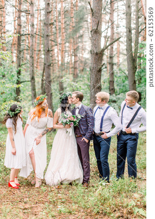 Beautiful kissing couple newlyweds with their friends having fun together. Happy bridesmaids and groomsmen with bride and groom on a walk in the forest 62153569