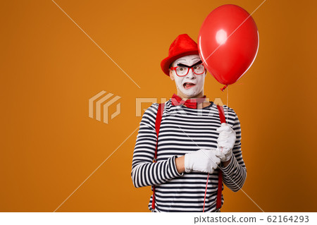 Sad mime man in vest and red hat with balloon in his hand on orange background Sad mime man in vest and red hat with balloon in his hand on orange background 62164293