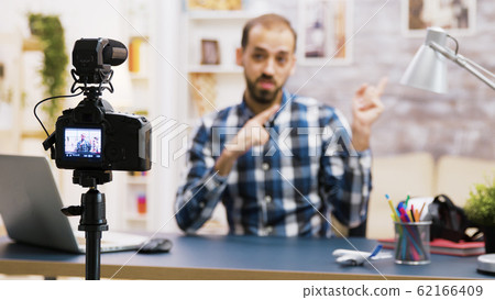 Vlogger sitting at desk in living room talking and looking at the camera Vlogger sitting at desk in living room talking and looking at the camera 62166409