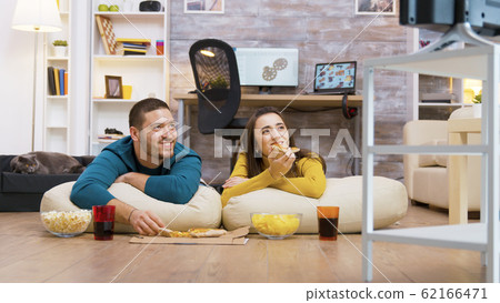 Happy caucasian couple eating pizza sitting on pillow Happy caucasian couple eating pizza sitting on pillow 62166471